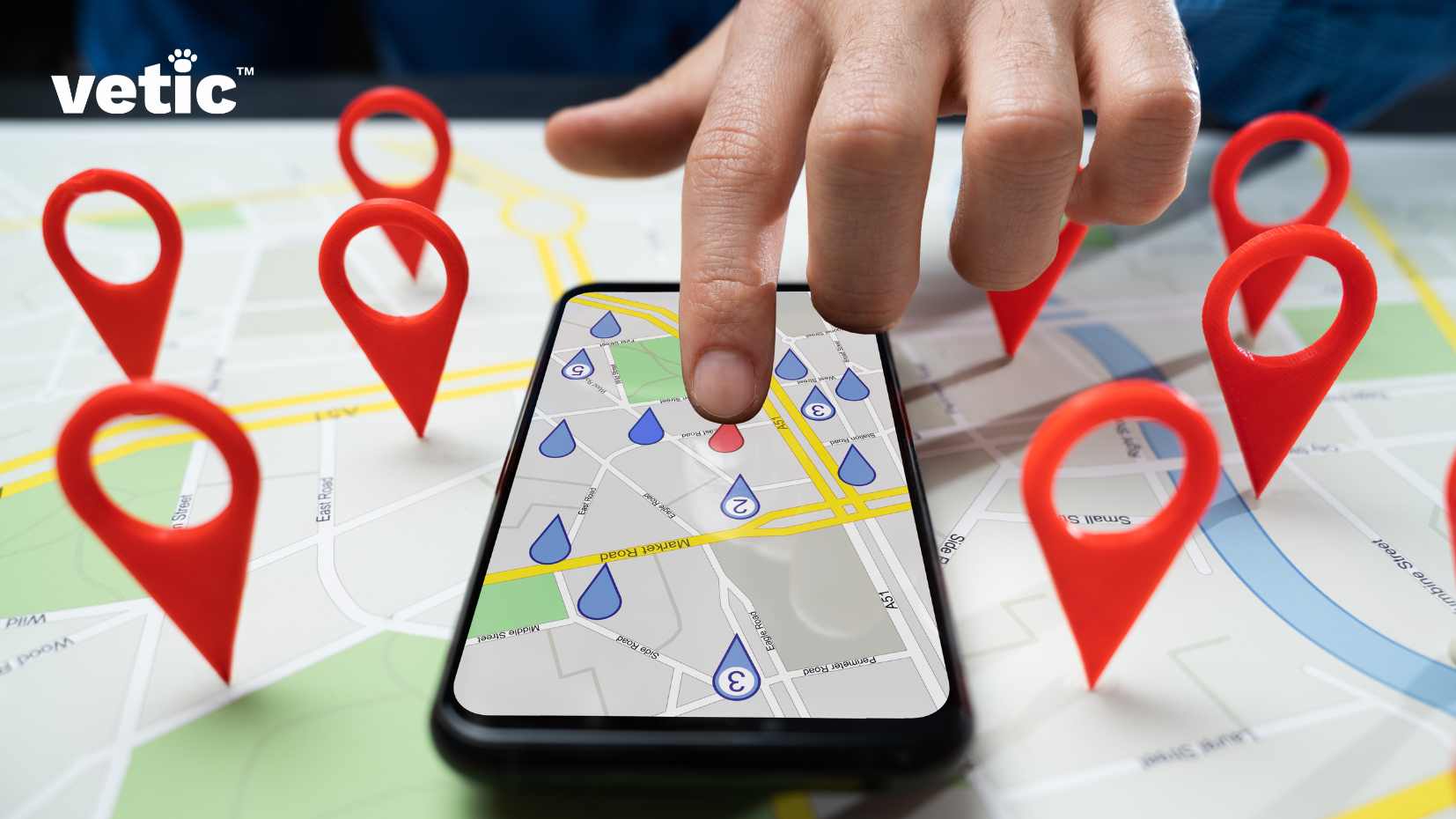 This image depicts a hand interacting with a smartphone that displays a map application. The map is populated with various location markers, and the background also shows an enlarged version of the same map. The central focus is on a hand, specifically a finger, tapping on a location on the smartphone. Search the best dog groomers near you on Google to help make a decision. The smartphone displays a digital map with streets and location markers. There are red location markers both on the phone’s screen and enlarged in the background, creating an effect of them popping out of the screen. The map displayed on the phone includes blue icons indicating specific places or services, like parking and information centers. In the top left corner, there is text displaying “vetic™”.